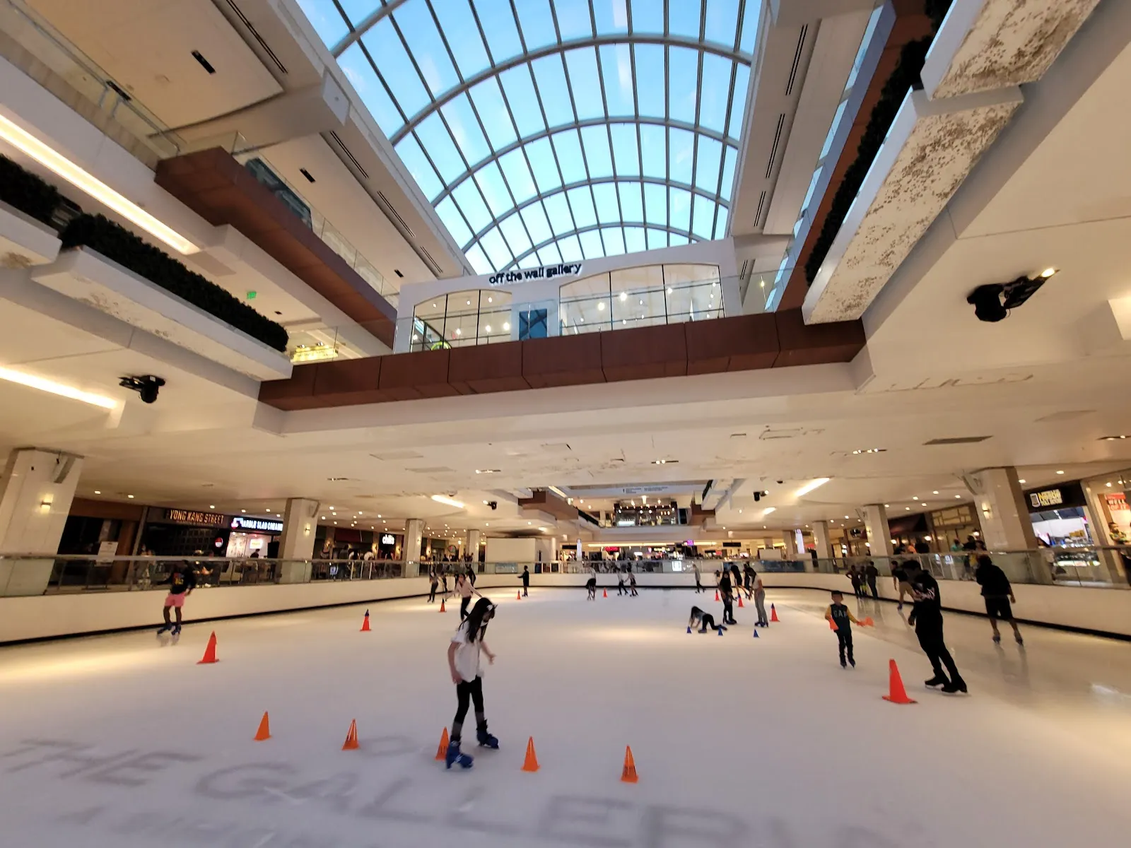 The Galleria's indoor rink under the long arched glass roof, skaters spread across the ice among orange cones with the mall's balconies and shops above. — Ice at the Galleria