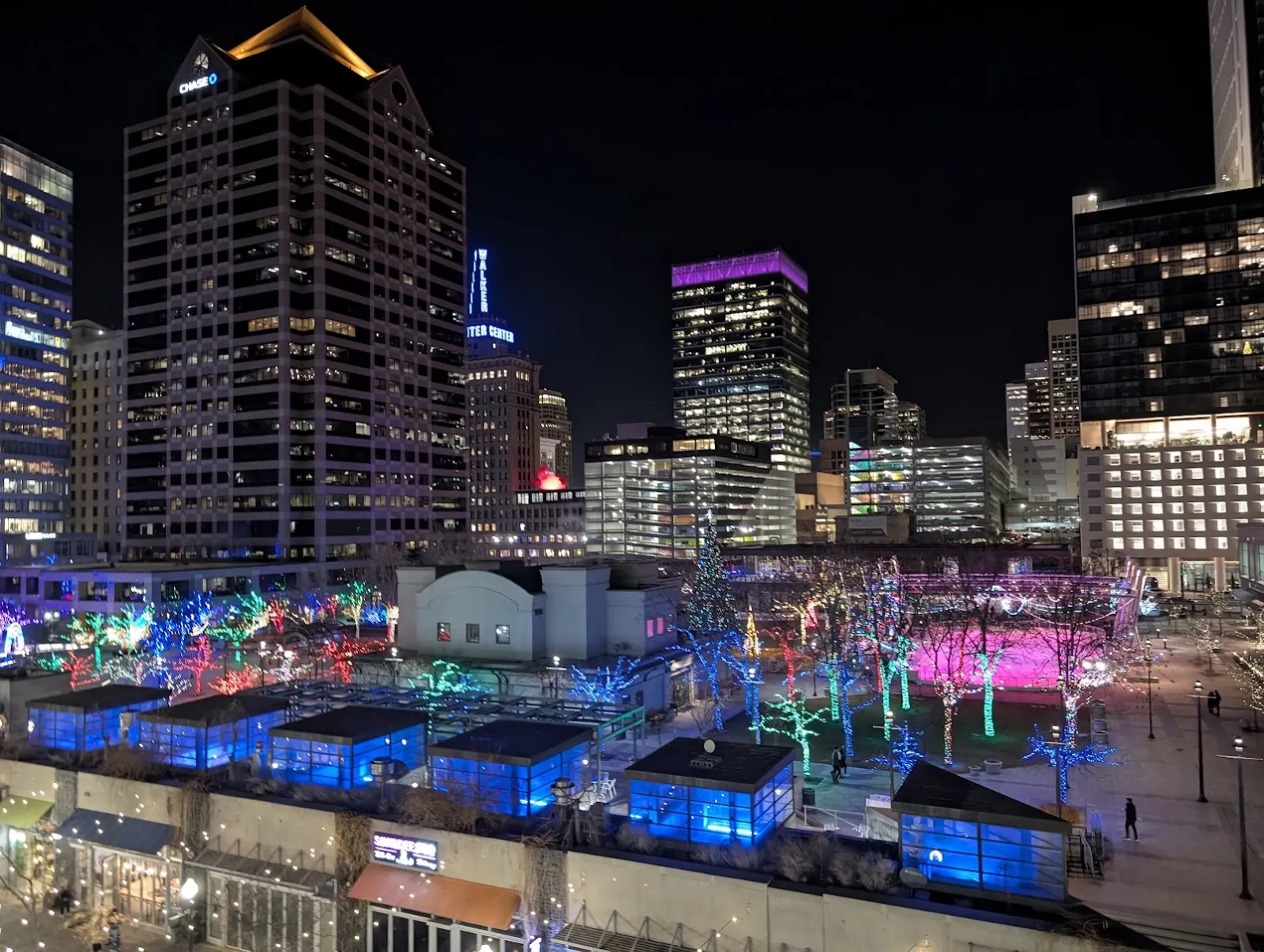 La plaza de noche desde arriba, con la zona de la pista, los pabellones iluminados en azul y las tiras de luces de colores en los árboles frente a las torres del centro. — Gallivan Center Ice Rink
