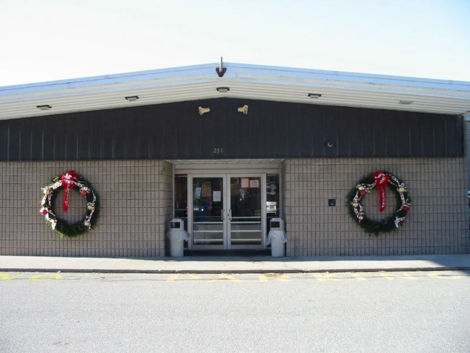 Front entrance of a low brick-and-metal building with a dark upper band, double glass doors under an address number 231, wreaths flanking the doors, empty parking lot in foreground — Gallo Ice Arena