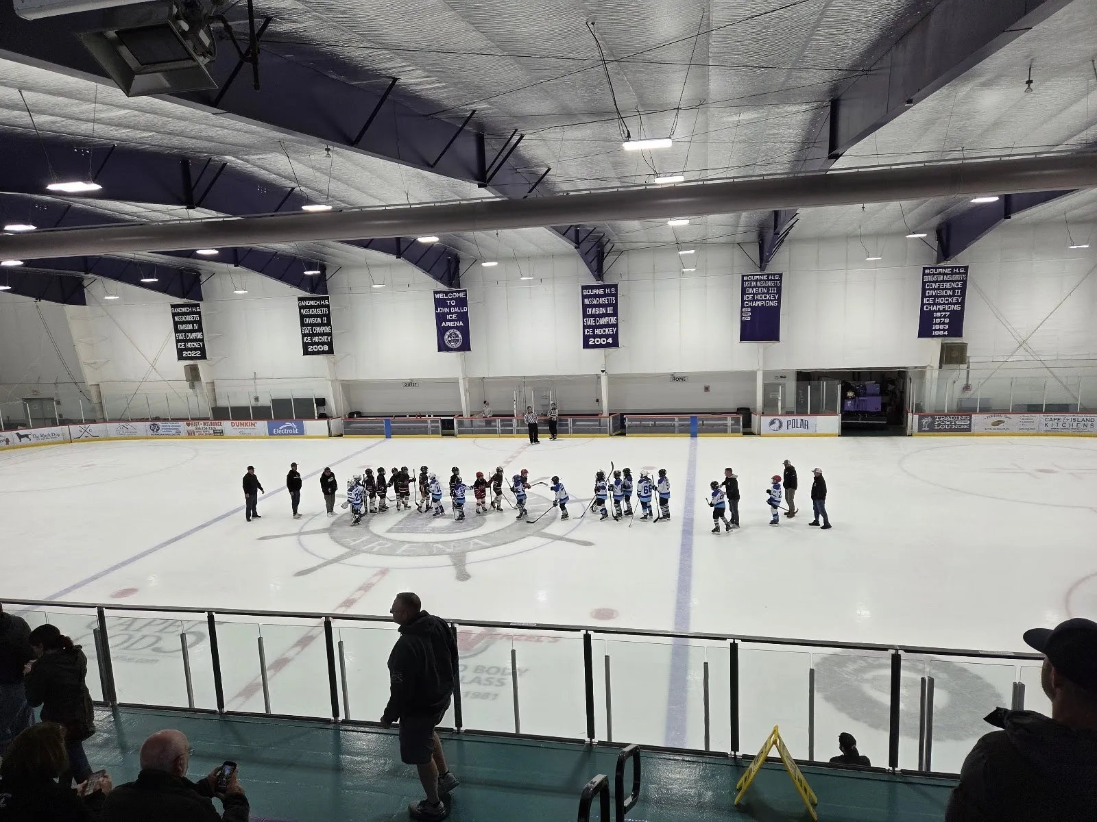 A wide view down the sheet from beside the stands, players spread across the ice beneath purple-braced steel beams with banners hung along the far wall. — Gallo Ice Arena