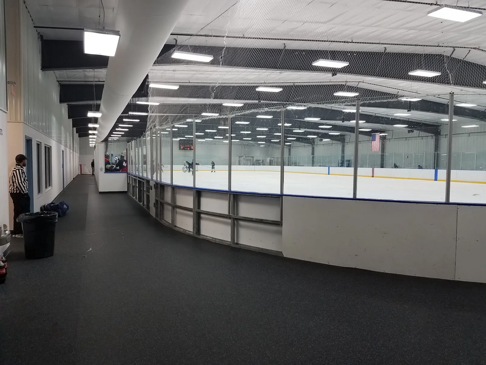 Parte de la pista vista desde el pasillo de espectadores junto a las vallas, con un árbitro y un partido de hockey en curso al fondo — The Garden Ice Arena