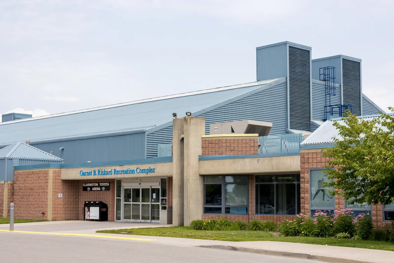 The Garnet B. Rickard Recreation Complex entrance, a brick front with the name above glass doors beneath tall blue-grey metal rink roofs. — Garnet B. Rickard Recreation Complex