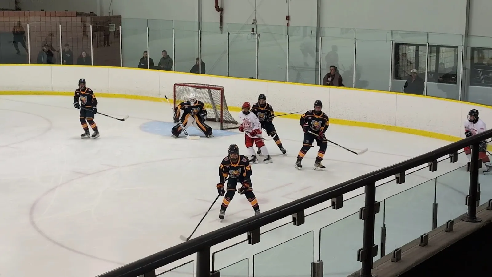 A hockey game on the ice seen from the viewing walkway behind glass: players in dark and white jerseys around a goal at the curved end boards, with the yellow-topped dasher and spectators along the far side. — Garnet B. Rickard Recreation Complex
