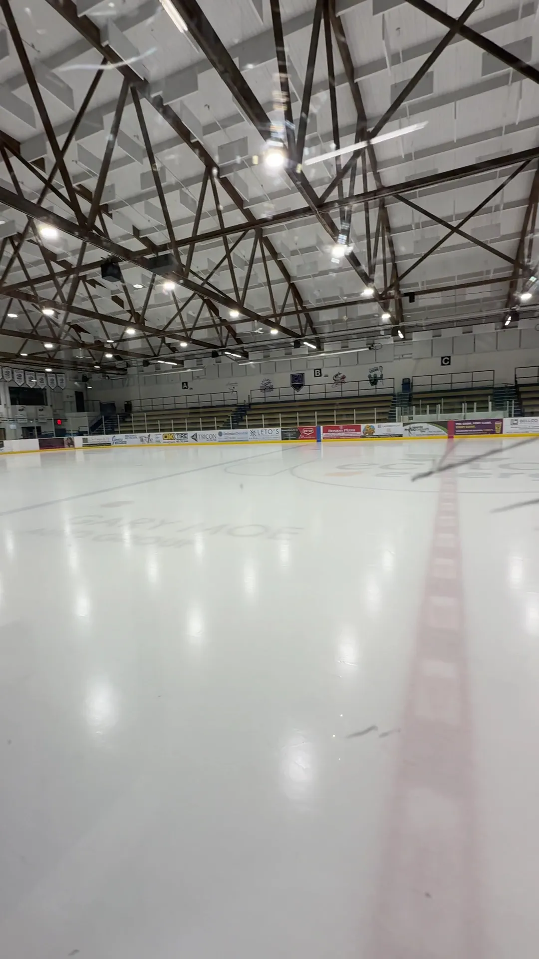 The empty ice seen from rink level through the glass, with the Gary Moe Auto Group logo at centre ice and the seating under a steel-truss roof. — Gary Moe Auto Group Sportsplex