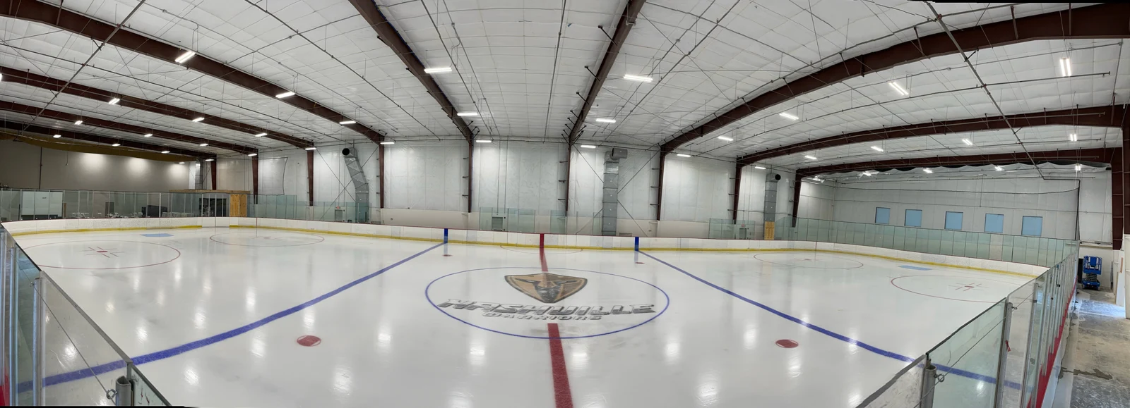 The full ice sheet and Warriors center-ice crest beneath exposed steel beams — Gary Force Acura Ice Arena