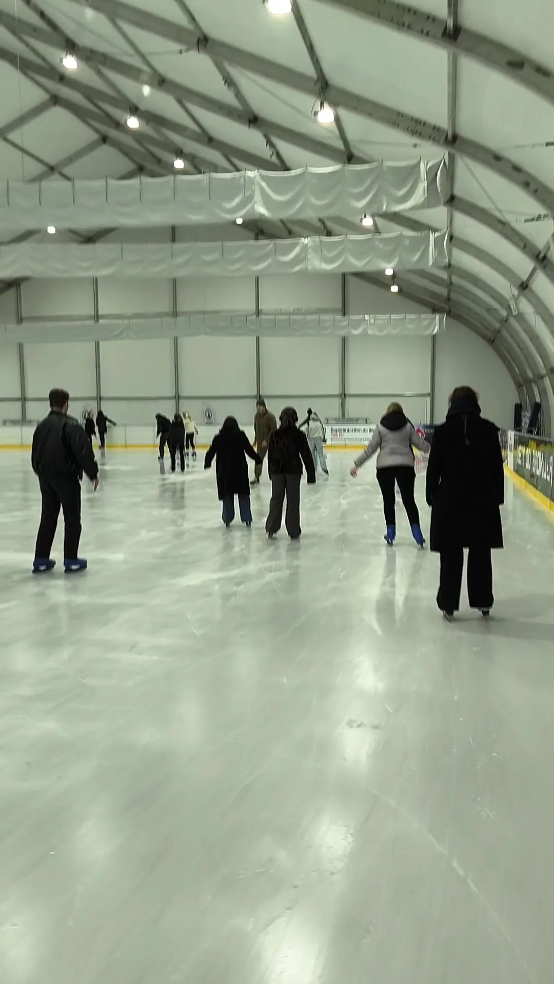 Skaters seen from behind on the ice inside the new tent hall at Plac Zebrań Ludowych, under its white fabric roof and steel frame. — Gdańskie Lodowisko (Plac Zebrań Ludowych)