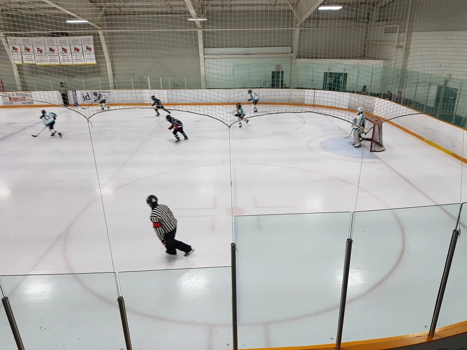 Players spread across a sheet seen from the glass, a faceoff circle in the foreground with a referee and a net at the far end under a long white roof. — Gemini Sportsplex (Strathroy)