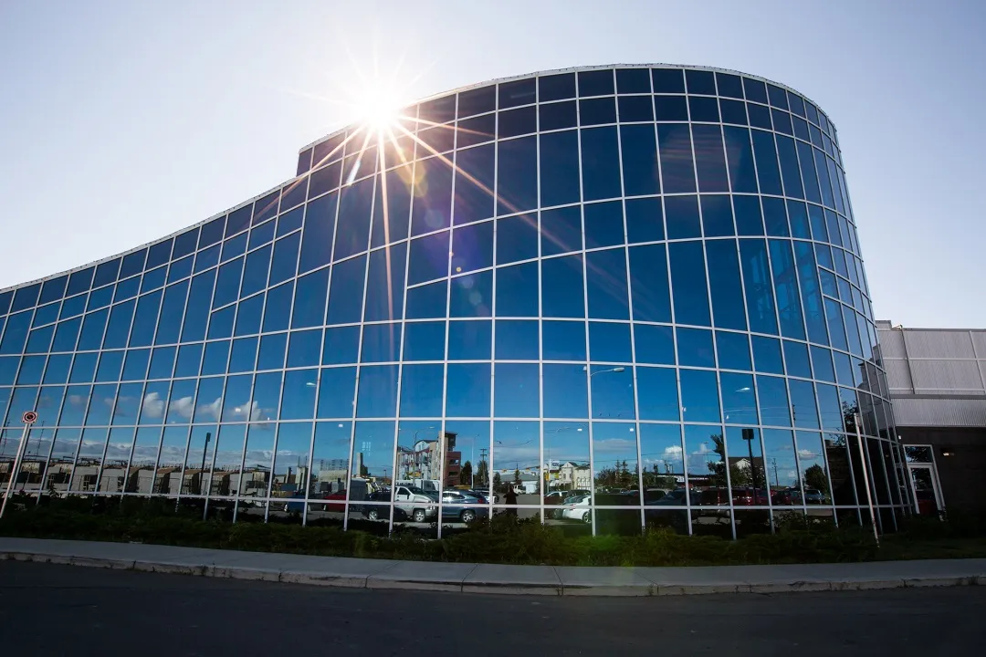 The curved glass facade of the Genesis Place building in the sun, with the lower roofline and the parking area in front. — Genesis Place Twin Arena