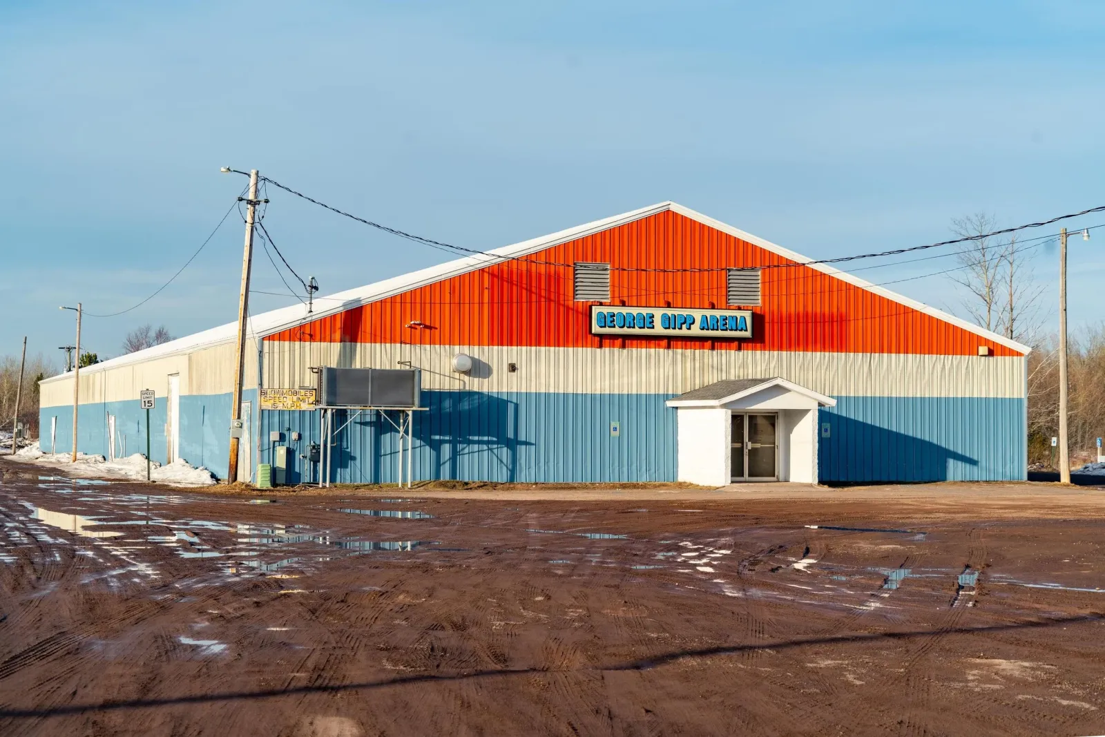 The George Gipp Arena: a long metal building painted red, white and blue with its name on a sign over a small white entrance porch, seen across a muddy lot in spring. — George Gipp Arena