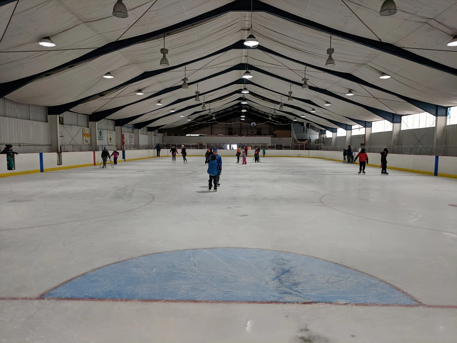 The ice sheet seen from the end of the arena, with a blue semicircle crease painted on the ice, skaters spread along the far side and a pitched roof of white fabric over steel trusses. — George Gipp Arena