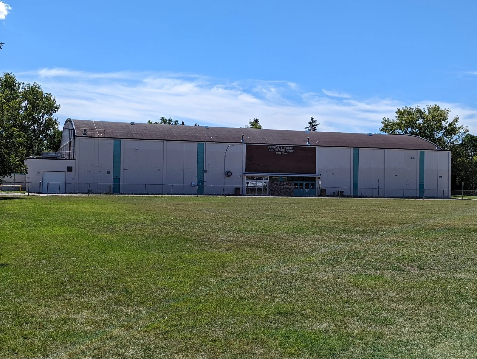 The long white George S Hughes South Side Arena in Edmonton, seen across the park lawn with its name sign over the entrance — George S Hughes Southside Arena