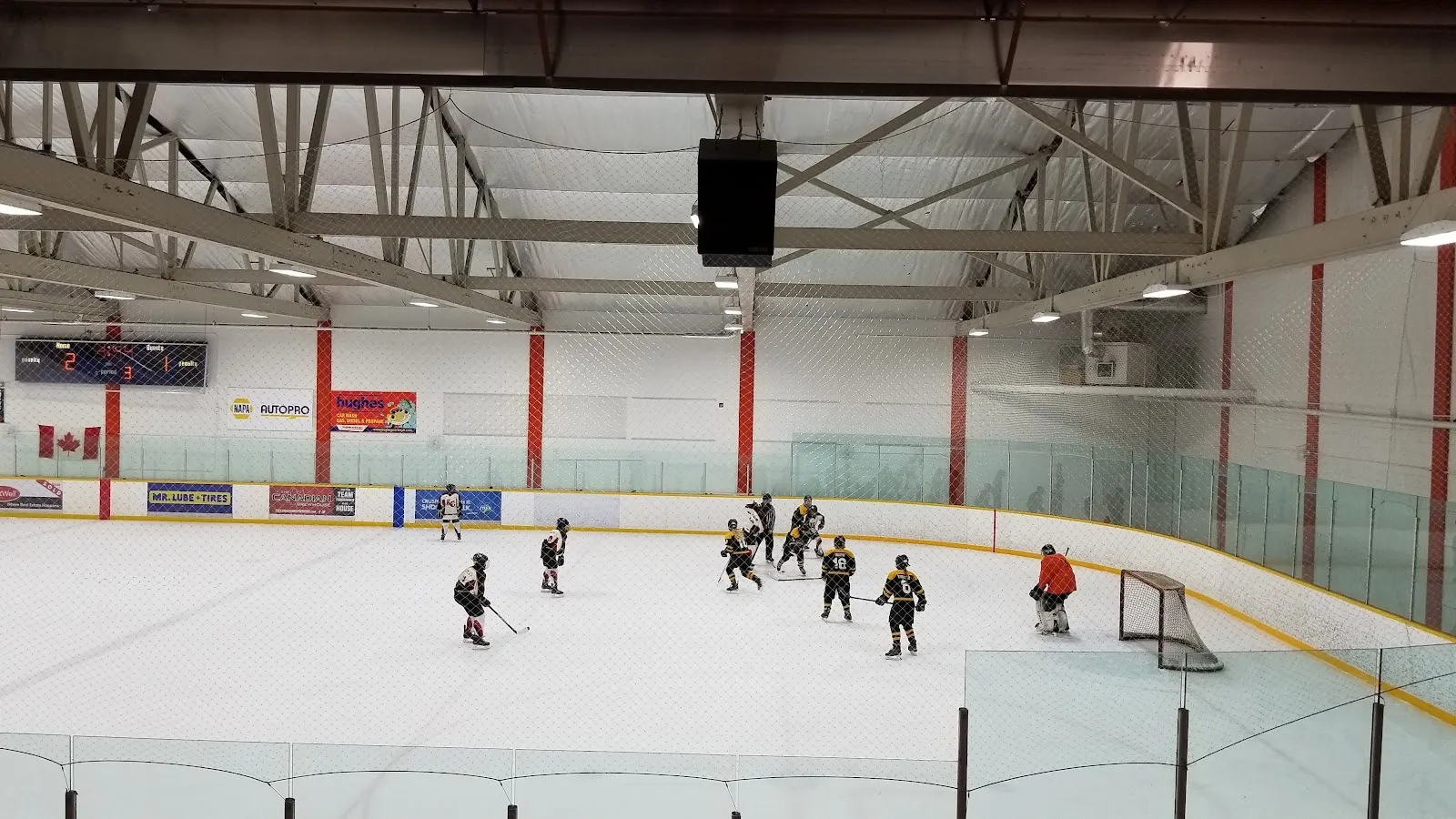 A game seen from the stands, players spread across the sheet beneath steel girders and red posts with sponsor boards along the far wall. — George S Hughes Southside Arena