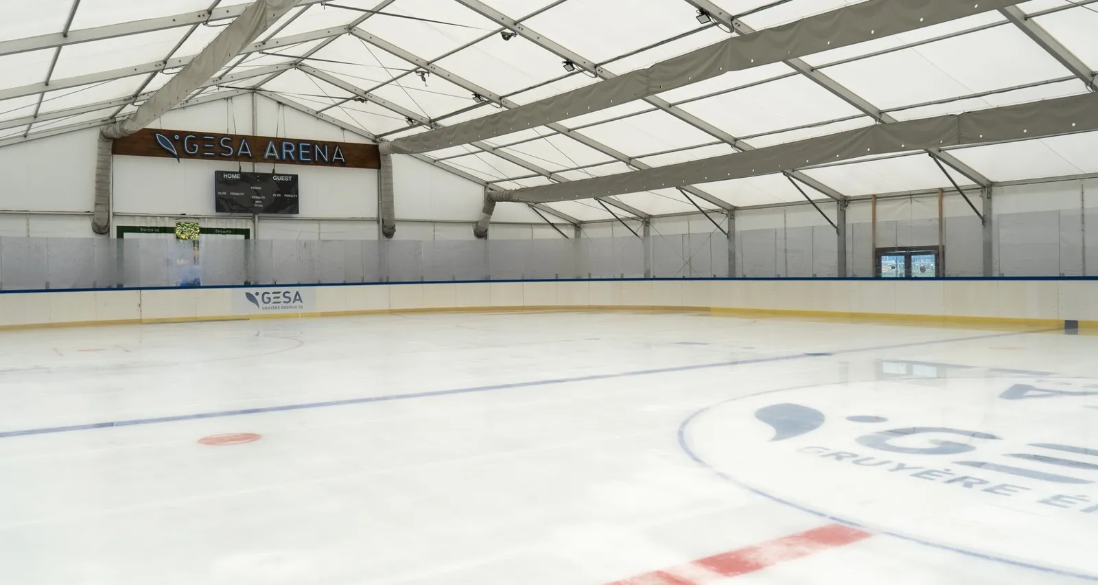 The ice surface of the GESA Arena in Charmey under its open roof frame, with the boards and the centre-ice logo. — GESA Arena