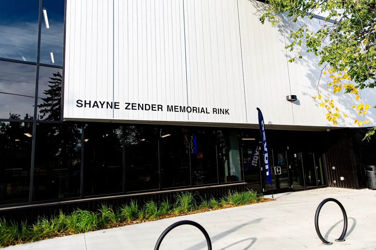 The glass entrance of the GFL Twin Arena in Devon, with the name Shayne Zender Memorial Rink in black letters on the white metal facade above the dark glazing, a bike rack and landscaping in front. — GFL Twin Arena