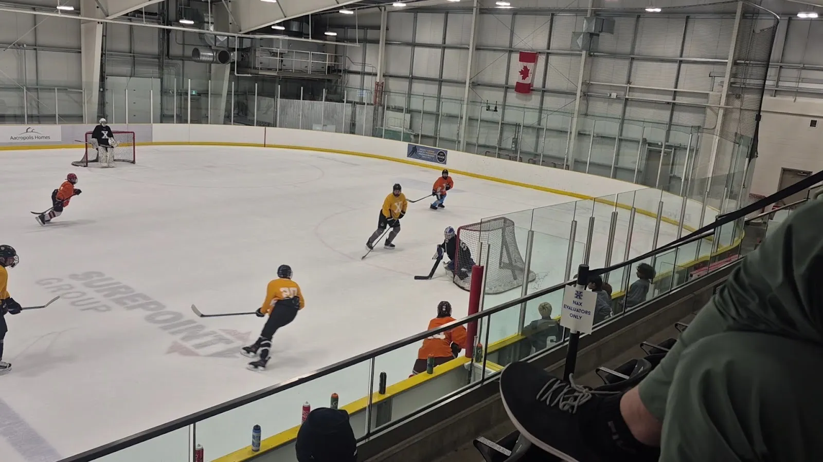 A game seen from the stands through the glass, players in yellow and red spread across the sheet with Canadian flags on the end wall. — GFL Twin Arena