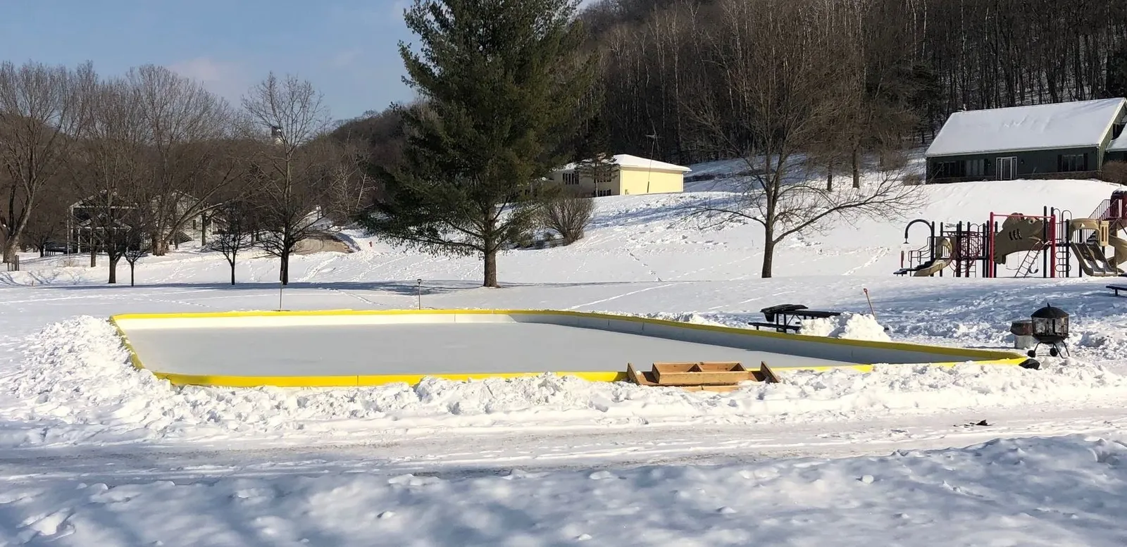 La pista de Jaybee Drive en Gilmore Valley, Winona: una superficie de hielo con borde amarillo sobre un campo nevado, con una caseta de calefacción y juegos infantiles detrás. — Gilmore Valley Ice Rink