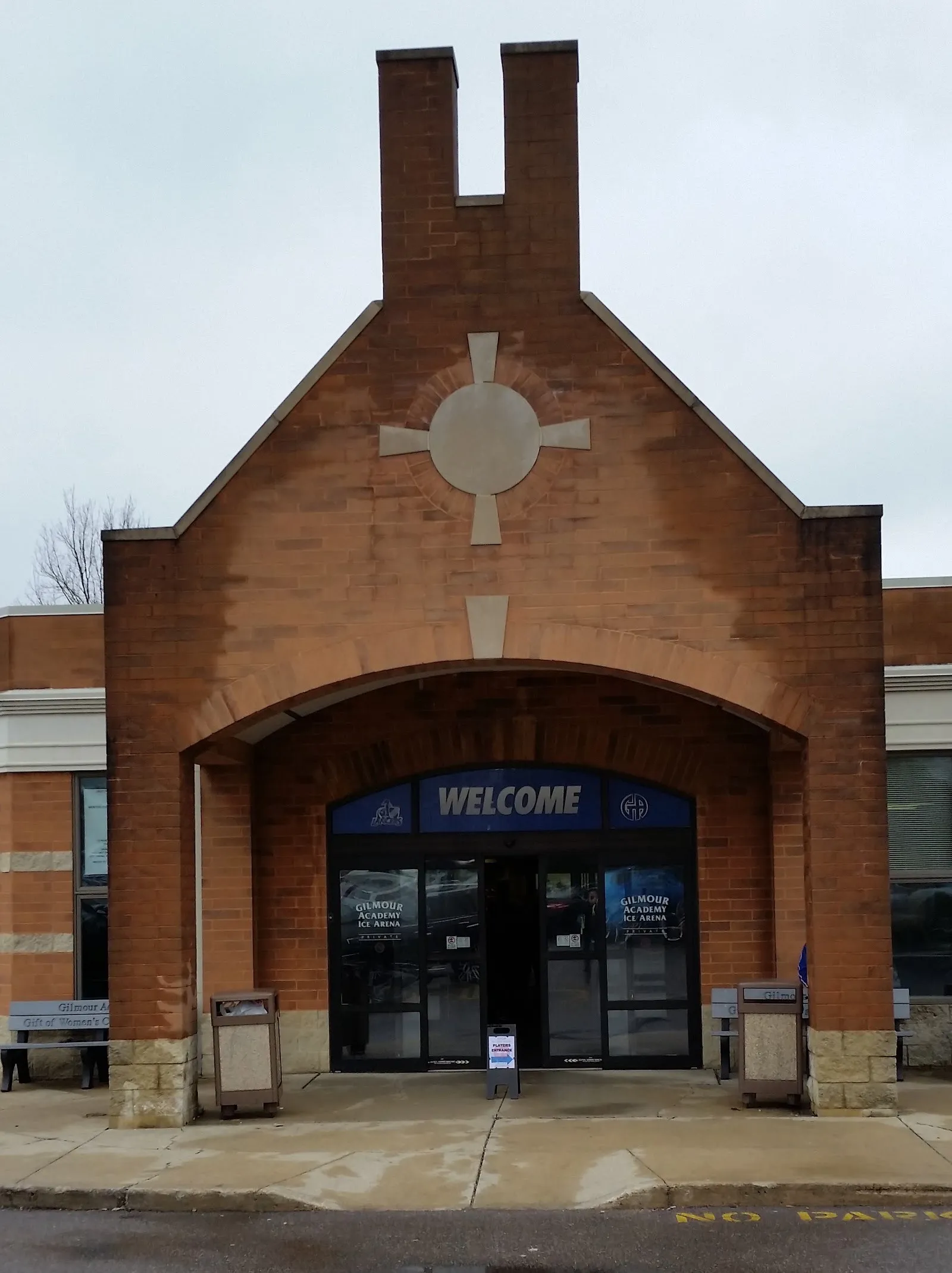 The academy ice arena entrance in Gates Mills: a brick gable with a stone cross over the arched porch and the blue Welcome sign above the doors — Floyd E. Stefanski Ice Center at Gilmour Academy