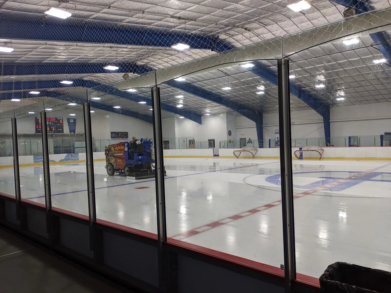 Empty NHL-sized ice sheet seen through the glass as a Zamboni resurfaces it, with a HOME/VISITOR scoreboard and a 'GA' logo at center ice. — Floyd E. Stefanski Ice Center at Gilmour Academy