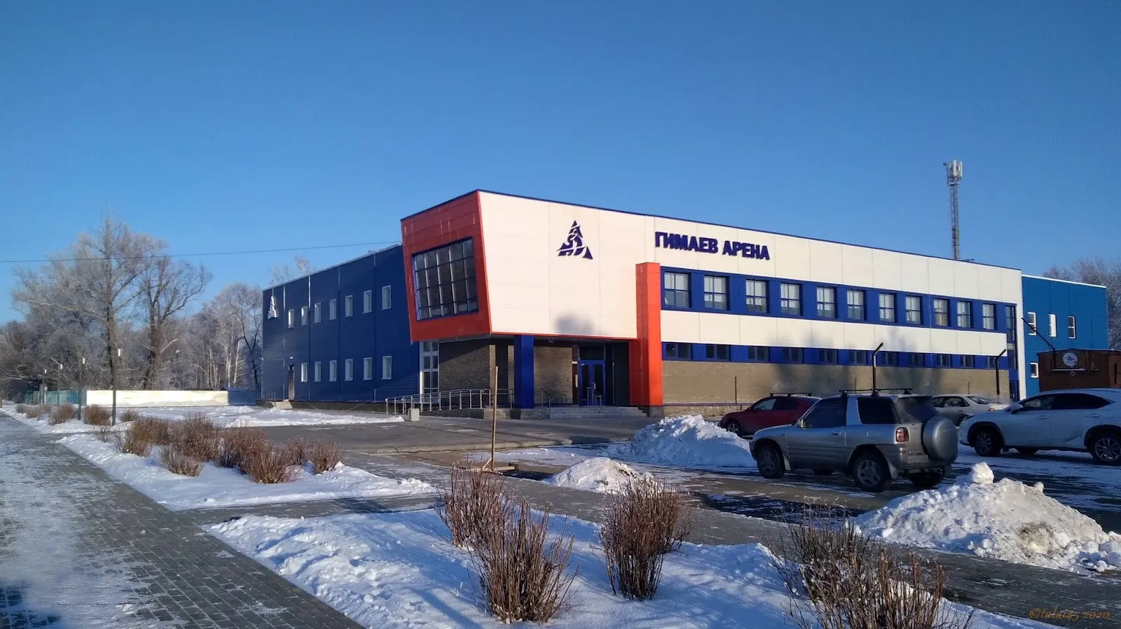 The Gimayev Arena ice palace in Rubtsovsk: a blue, white and orange building with the arena name over the entrance, in winter sunlight — Gimayev Arena Ice Rink Rubtsovsk