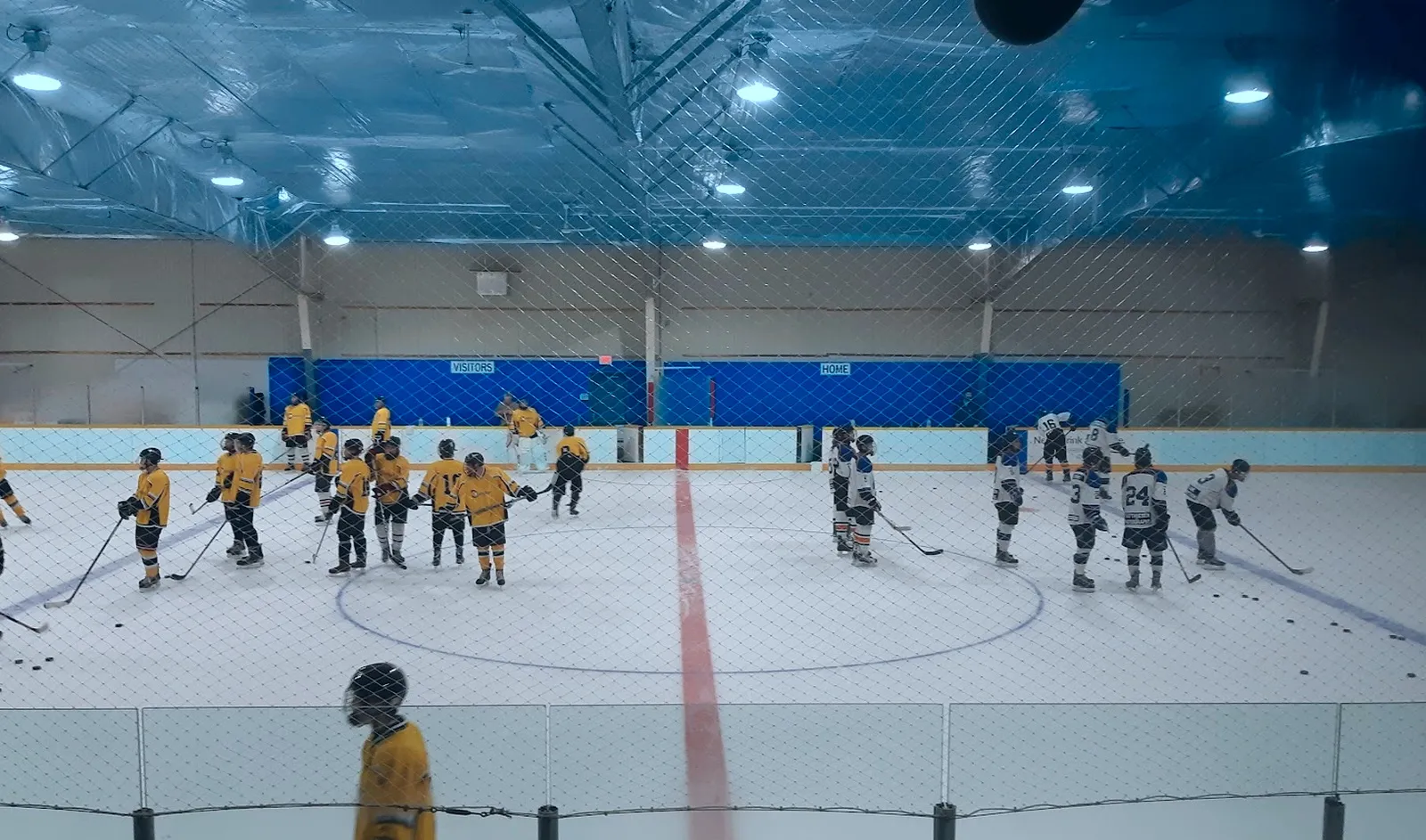 Players lined up along a red line on a wide sheet under a blue-lit ceiling, a goalie's helmet at the bottom edge of the frame. — Gimli Recreation Centre