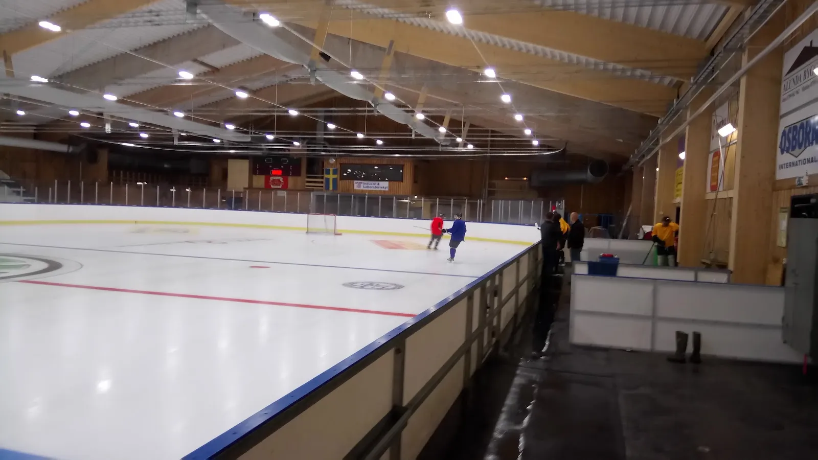 Isen sedd längs sargen från ena kortsidan av hallen, tom förutom ett par avlägsna åkare, under ett ljust tak av trä och stål med lysrör. – Gimo ishall