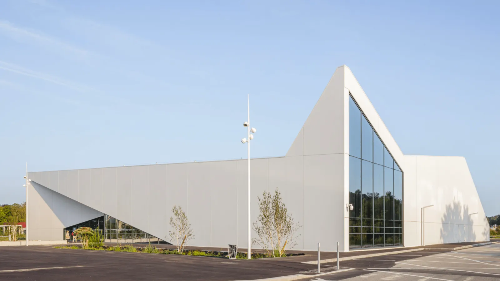 The building seen from the car park in low sun: white angular volumes with a tall pointed gable and a glazed end wall. — Glacéo