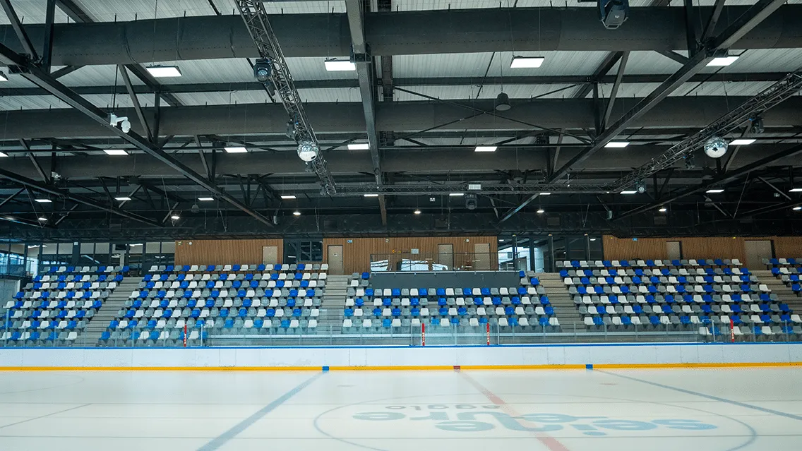 The empty ice with the Glacéo logo at centre, blue and white tip-up seating along one side and a steel-truss roof with disco balls above. — Glacéo