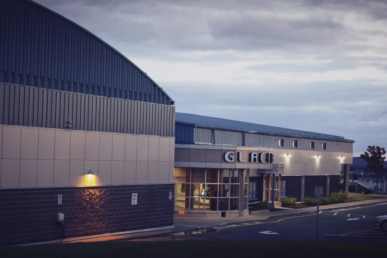 Exterior al atardecer del edificio de techo curvo de Glacier Arena, con el letrero iluminado de "GLACIER" sobre la entrada de vidrio y el estacionamiento en primer plano. — Glacier Arena