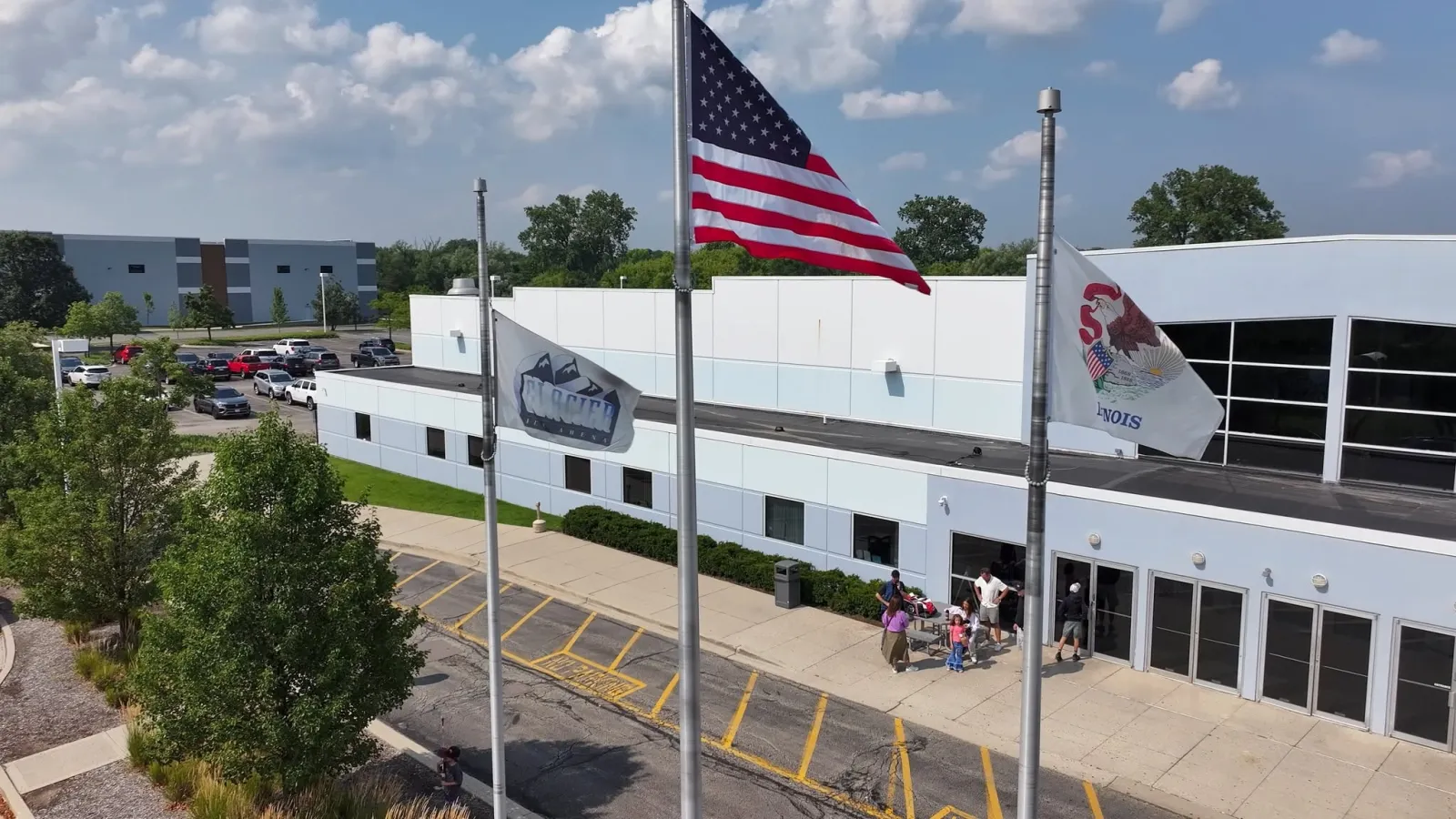 Aerial view of the building's front entrance and parking lot, with a "Glacier Ice Arena" flag flying alongside the US and Illinois flags — Glacier Ice Arena (Vernon Hills)