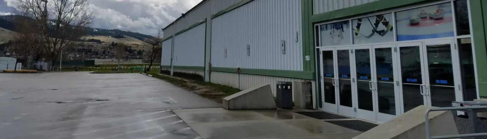 El muro exterior de metal corrugado verde y blanco de la pista y las puertas de vidrio de la entrada decoradas con gráficos de hockey y curling, vistos desde el otro lado de un estacionamiento vacío con montañas detrás. — Glacier Ice Rink