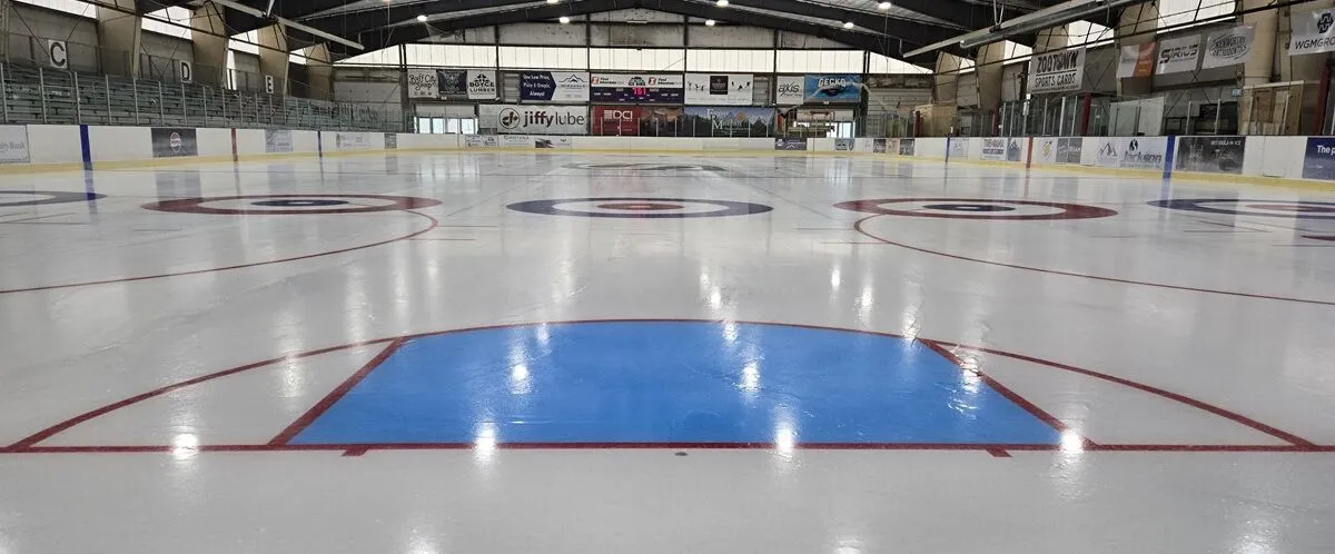Toda la pista vacía con las dianas de curling y un área de portería de hockey azul, vallas con mantas de patrocinadores (incluida una de Zootown Sports Cards) y armaduras en el techo. — Glacier Ice Rink
