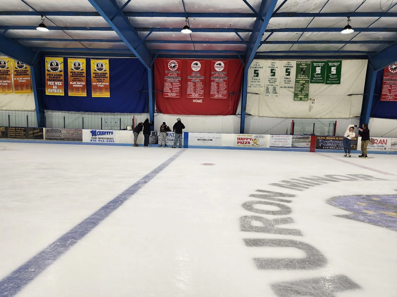 El hielo visto desde las vallas del extremo, con una línea azul y un logotipo en el borde, y banderines colgados de las vigas azul oscuro del techo sobre la pared del fondo. — Glacier Pointe Ice Complex