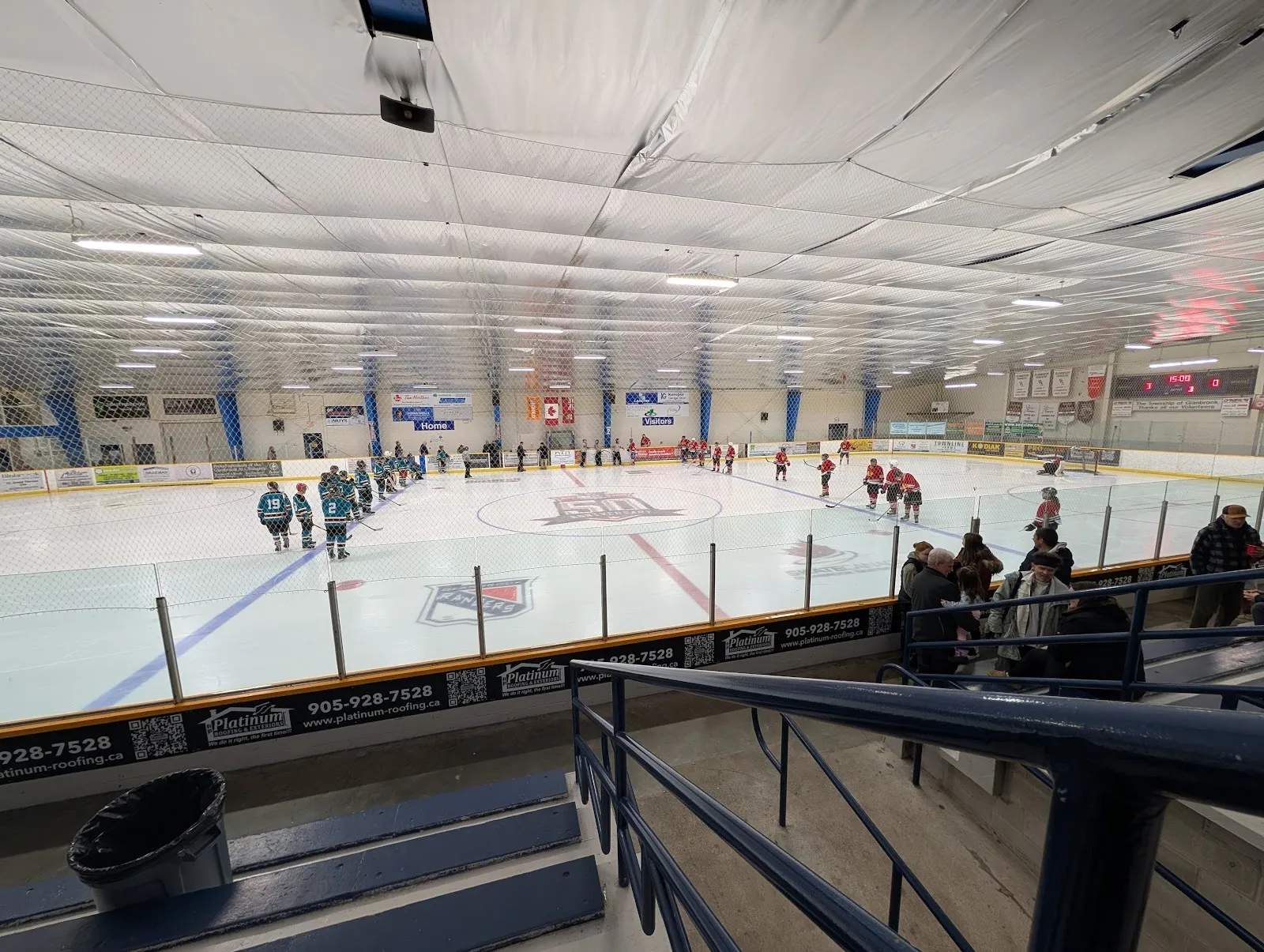 A wide view of the sheet from the stands, players spread across the ice under a long grey ceiling with a steel-trussed roof. — Glanbrook Arena