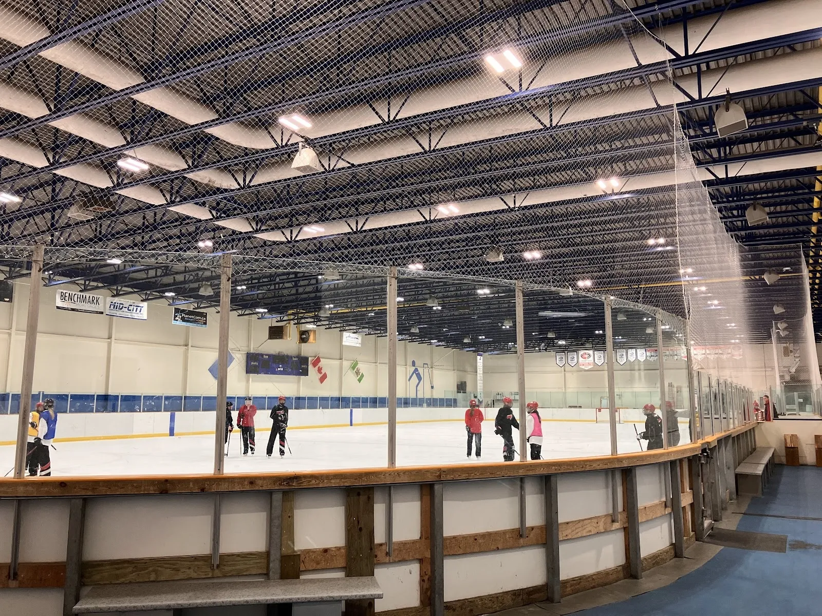 Una pista amplia vista a lo largo de un barandal de madera, con patinadores repartidos por el hielo bajo un oscuro techo de acero con una franja azul de vallas. — Glen Allan Recreation Complex