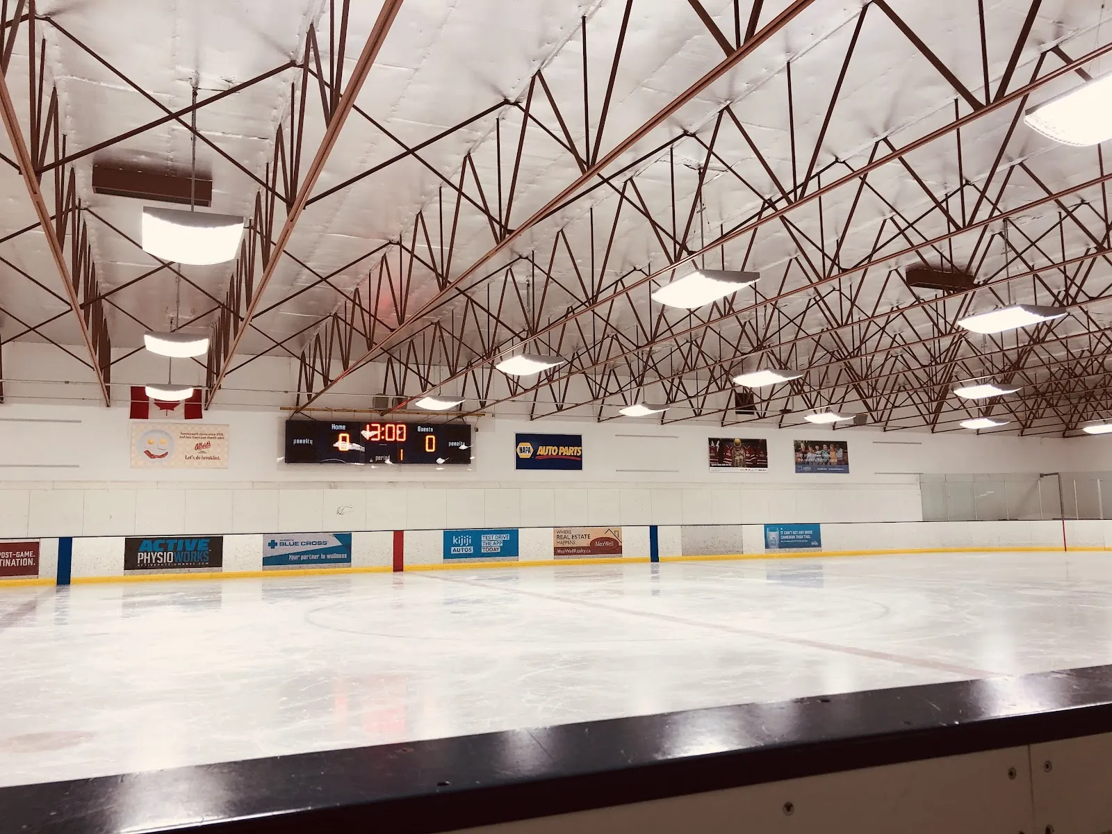 Empty ice rink from the boards showing the full sheet, a scoreboard reading 0-0, a Canadian flag and rink-board sponsor ads (NAPA Auto Parts, Kijiji Autos, MaxWell Realty) along the far wall, exposed steel roof trusses overhead. — Glengarry Arena