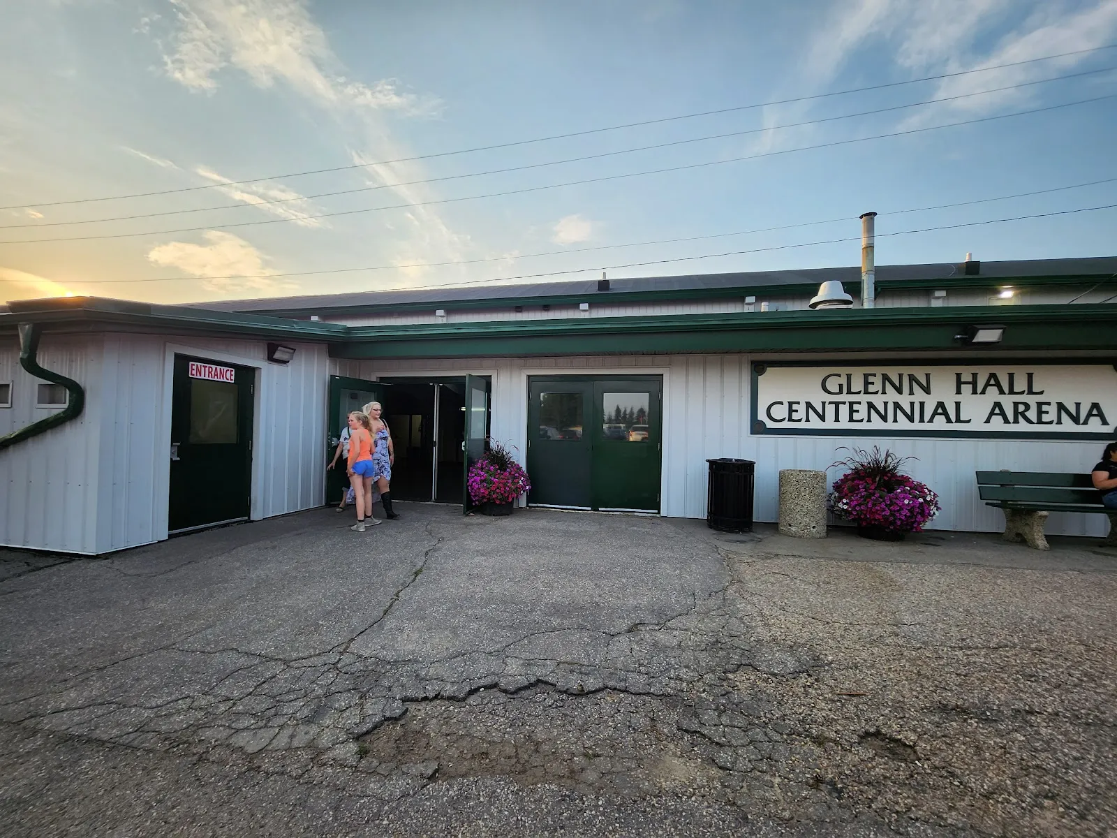 La fachada del Glenn Hall Centennial Arena al anochecer: un edificio bajo y blanco con puertas verdes, el nombre del arena en un cartel sobre la entrada y una explanada pavimentada con macetas de flores. — Glenn Hall Centennial Arena