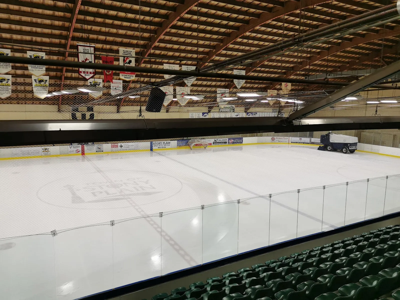 El hielo de Glenn Hall Centennial Arena visto desde los asientos verdes, con mantas de campeonato colgando bajo las armaduras de madera del techo. — Glenn Hall Centennial Arena