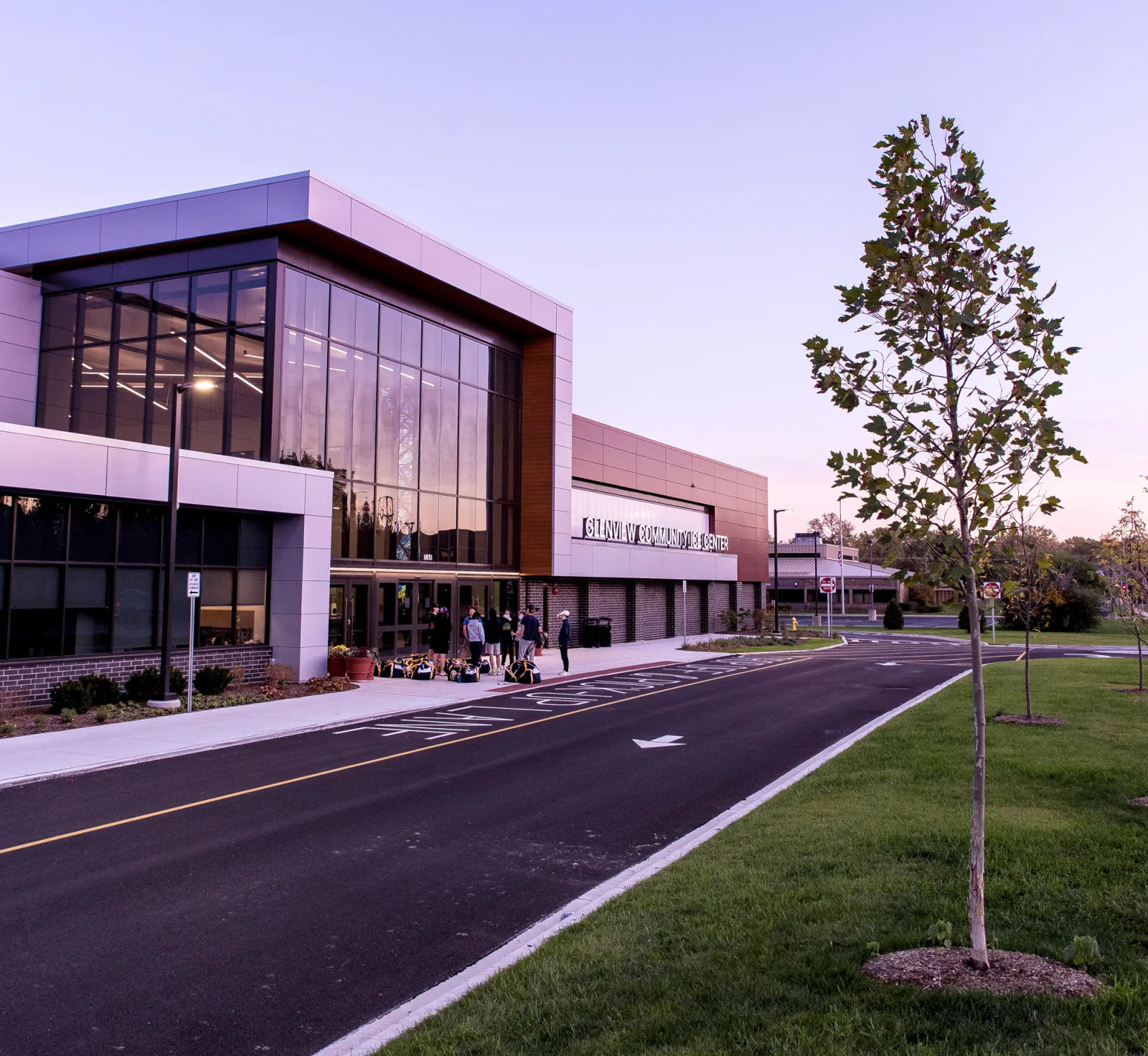 The front of the ice center at dusk: a glass-walled two-storey entrance with wood-panel trim, "GLENVIEW COMMUNITY ICE CENTER" lettering along the façade, and a few people with hockey bags at the doors beside the drop-off lane. — Glenview Community Ice Center