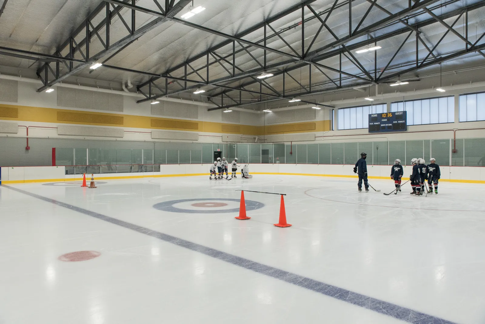 A full NHL-sized sheet from ice level with faceoff circles and blue lines in view, a group of young players in a lesson by the far boards, orange cones on the ice, a scoreboard on the end wall and a band of daylight windows above the boards. — Glenview Community Ice Center