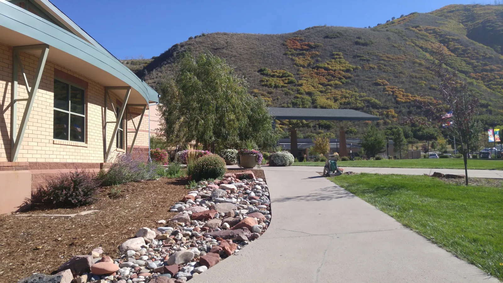El edificio color arena del centro comunitario junto a una jardinera de piedras y un andador, con pasto y la ladera de la montaña detrás en un día soleado. — Glenwood Springs Community Center Ice Rink
