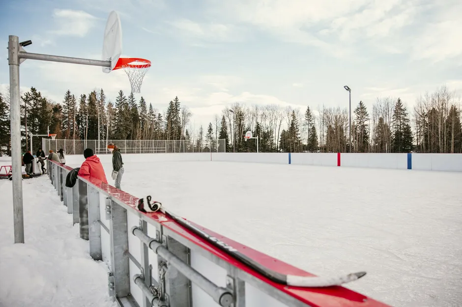 A lo largo de las vallas rojas de la pista exterior de Glenwood Park en invierno, con un aro de baloncesto sobre las vallas, algunas personas a lo lejos y abetos detrás. — Glenwood Park Outdoor Arena (Edson)