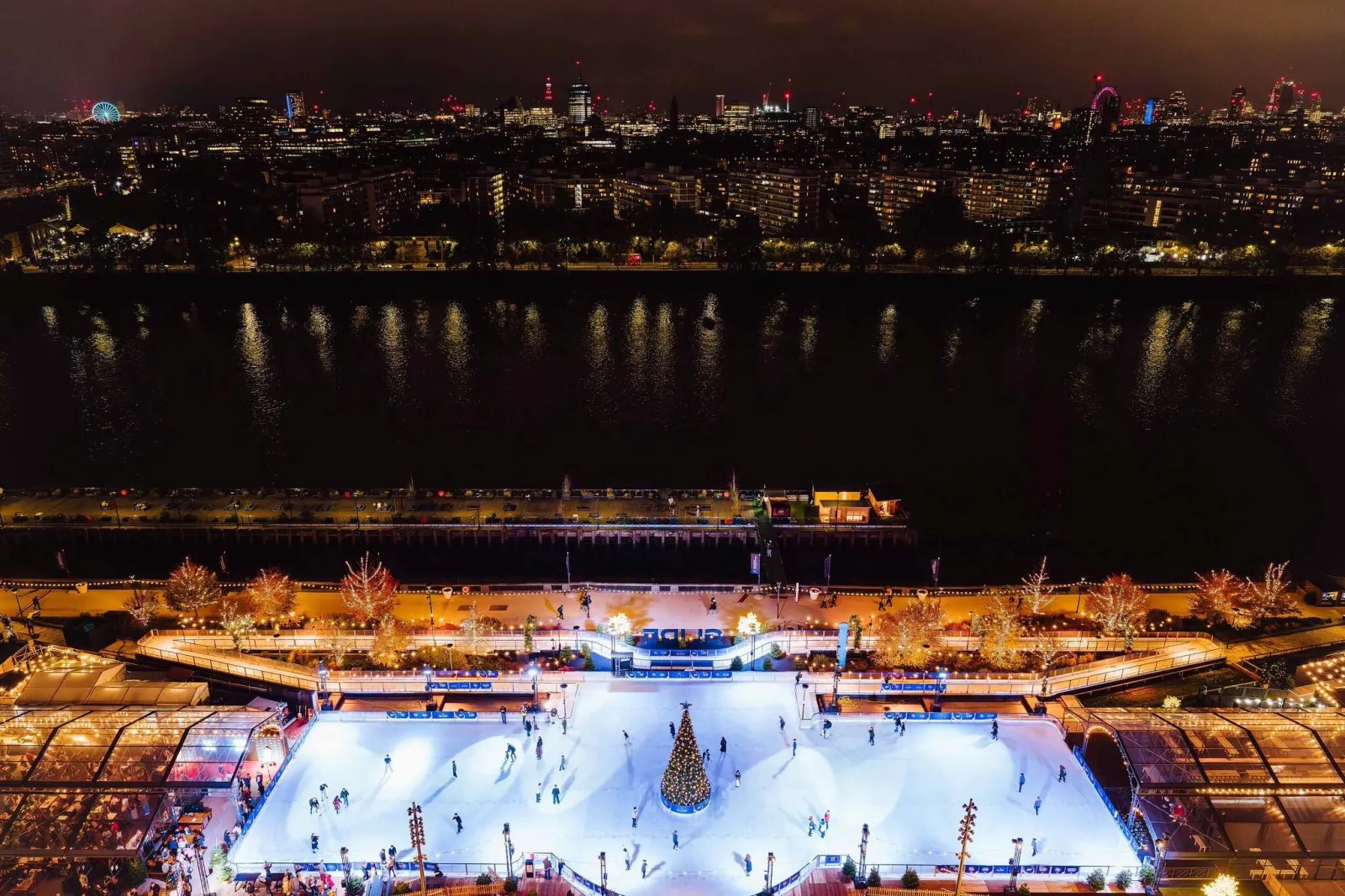 Dusk aerial of the Glide rink beside the river at Battersea Power Station, the white ice lit by the tree and surrounded by the lit terrace; skaters are small. — Glide at Battersea Power Station