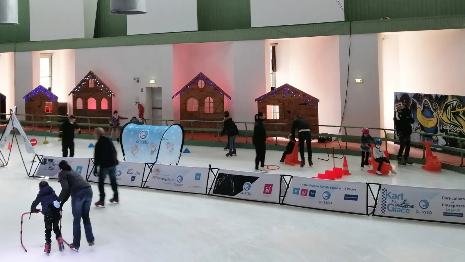 La patinoire couverte de Glisséo Cholet aménagée avec de petites maisons de style chalet le long des bandes, avec des patineurs et des enfants sur la glace. — Glisséo Patinoires