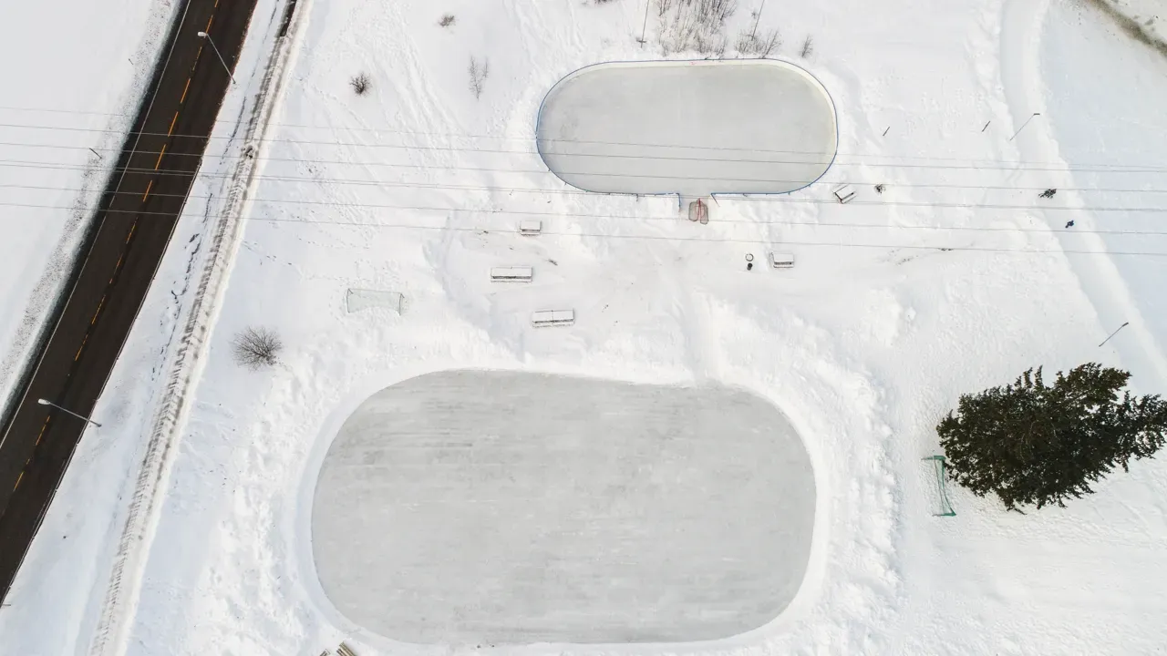 An aerial view of the two rounded ice rinks at Glostrupdumpa in winter snow, with a road running past on the left. — Glostrupbanen skøytebane (Lillestrøm)