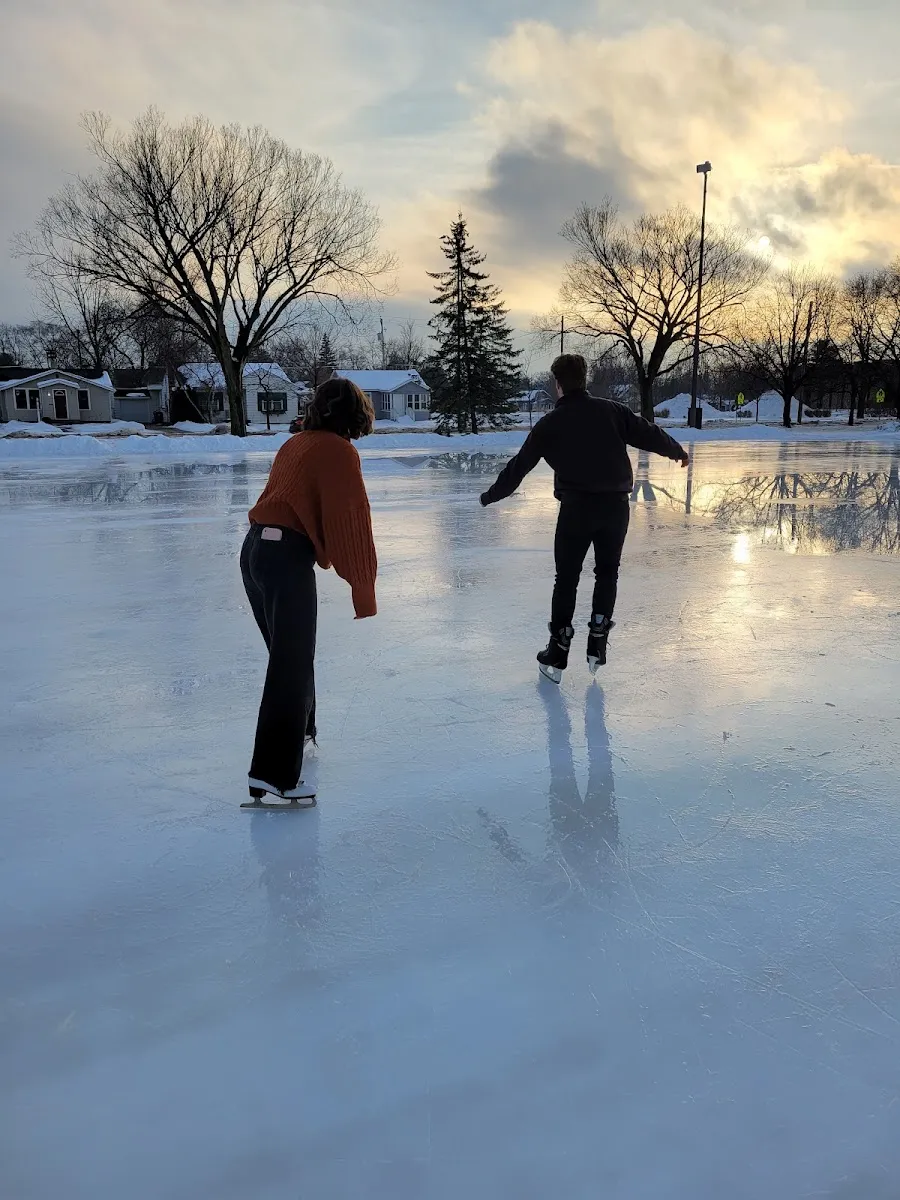 Two skaters on the outdoor rink ice at dusk, with houses and trees beyond — Goerke Park Ice Rinks
