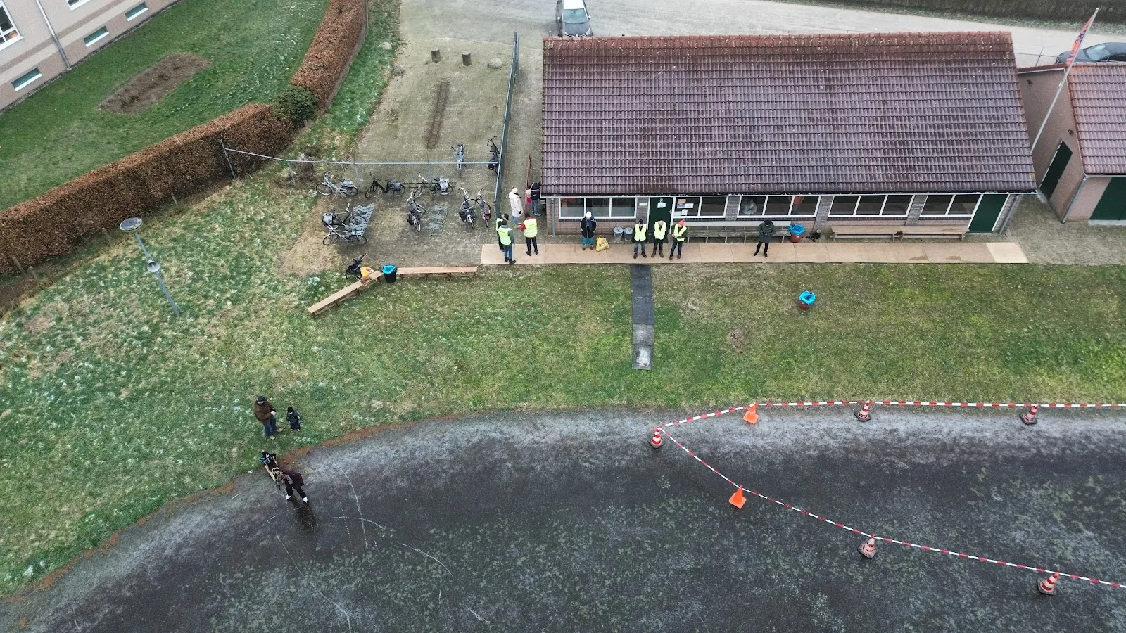 An aerial view of the Goor natural-ice rink beside the club's long shed roof, with skaters tiny on the ice. — IJsbaan 't Slag (Goorse IJsclub, Goor)
