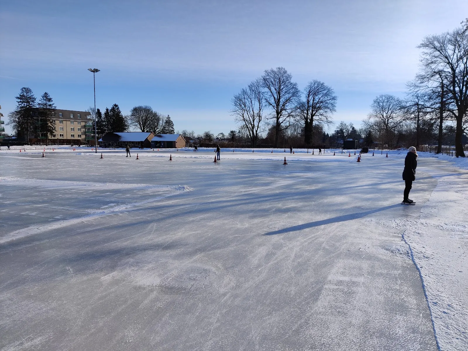 The grey natural-ice surface of the Goor club rink with a single skater in the distance and houses along the edge. — IJsbaan 't Slag (Goorse IJsclub, Goor)