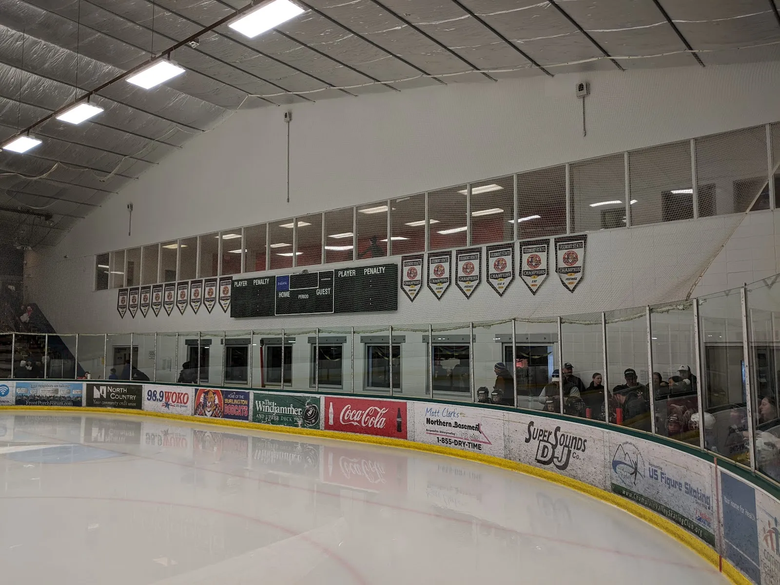 Vista junto a la pista de la superficie de hielo y las vallas cubiertas de anuncios de patrocinadores locales, con mantas de Vermont State Champions y un marcador en la pared de arriba, y espectadores visibles detrás del cristal. — Gordon H. Paquette Ice Arena