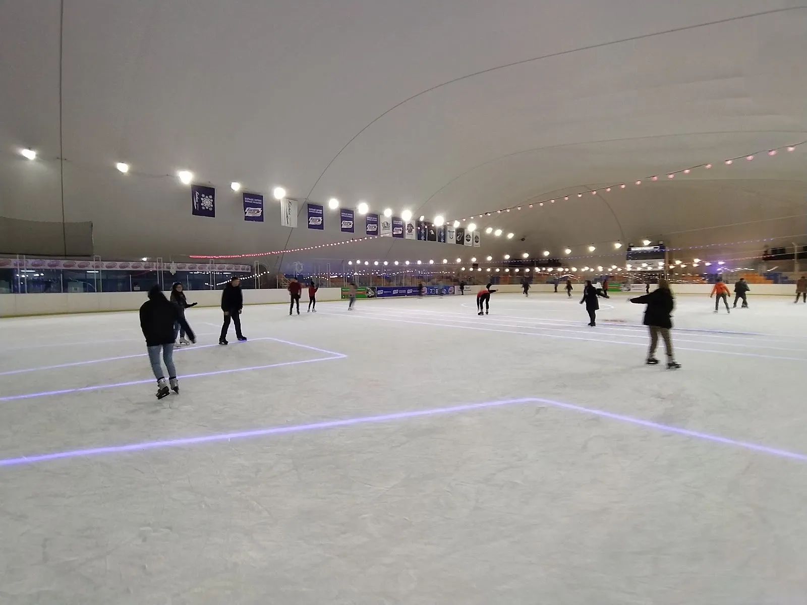 The ice sheet of the Bishkek city rink under its arched roof, with a few skaters in the distance — Gorodskoy Katok (Nomad Arena)