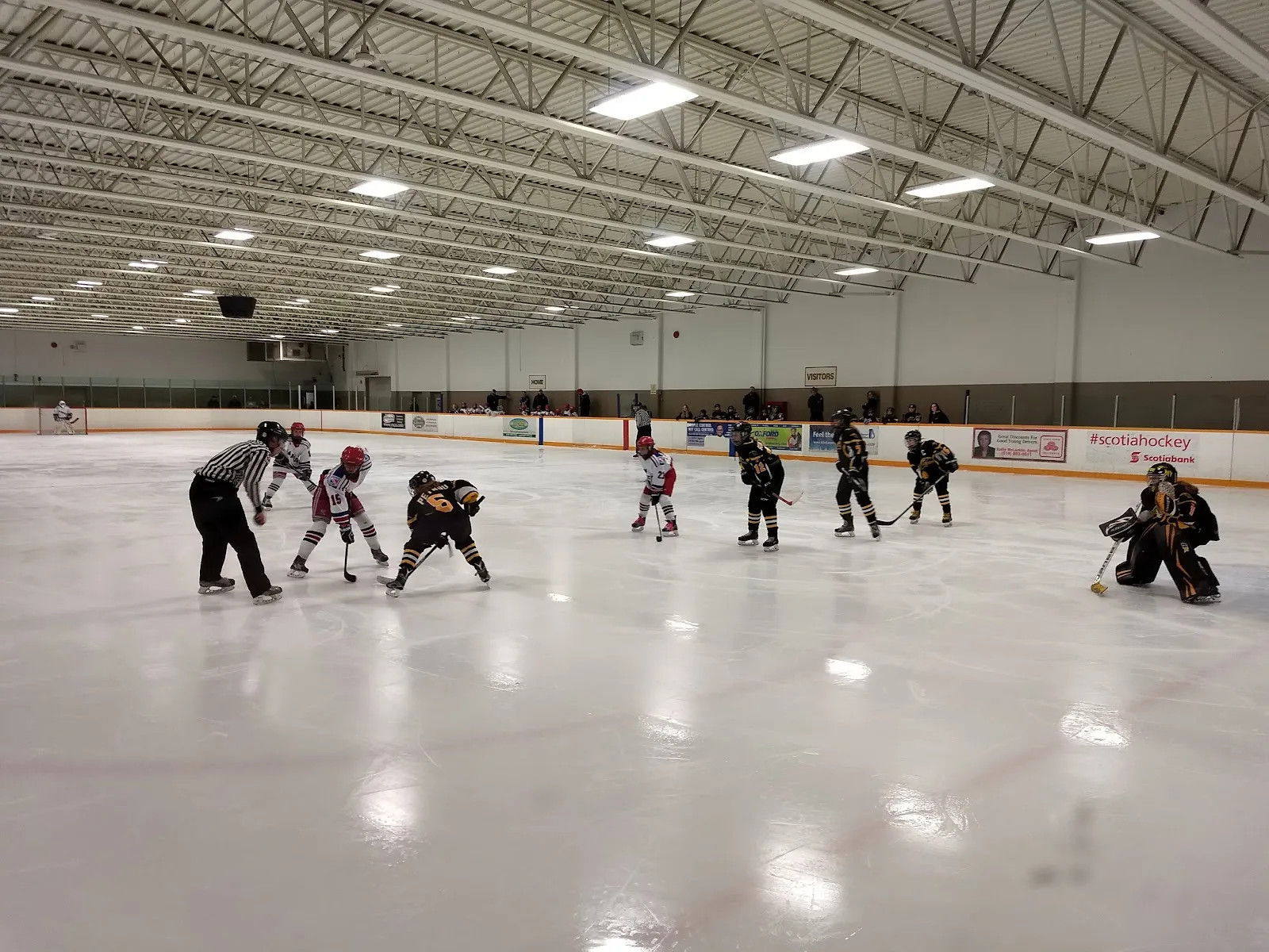 Un saque inicial de hockey juvenil en el extremo lejano del hielo, visto de lado bajo las cerchas del techo del estadio. — Grand River Arena